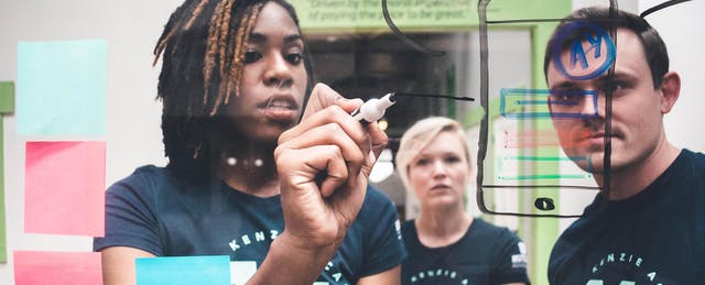 People in Kenzie Academy shirts sketching and arranging sticky notes on a glass writing wall